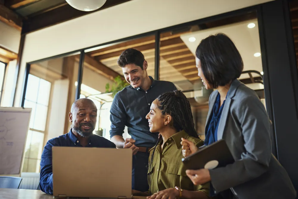 ALT Text: Project manager and teammates smiling cheerfully at team meeting in boardroom, excited over business plans on laptop